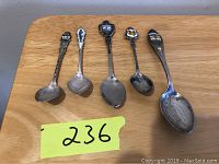 Photo showing five miniature sterling silver souvenir spoons laid out on a wooden surface, each with colorful enamel decoration on the handle.