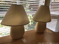 Two matching ceramic base table lamps with beige pleated shades sitting on a wooden surface near window blinds.
