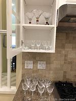 Set of glassware displayed on kitchen shelf and countertop showing variety of glass types including cut crystal glasses and martini glasses with red decoration on stems.
