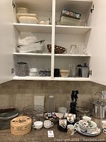 Photo of three shelves and countertop showing assortment of ceramic, glass, and plastic kitchenware including Brie Baker in box, Mikasa bowls, mugs, plates, measuring cups, storage containers, and decorative items