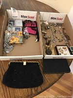 Wide view showing two boxes holding assorted jewelry and accessories plus two black evening bags laid on table.