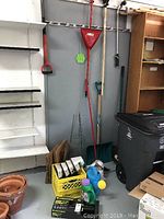 Photo showing assorted garden tools hanging on a wall and placed on floor including shovels, rake, pots, hand tools, hose and sprinkler box
