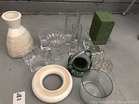 Wide shot showing all vases and bowls on floor including ceramic vase, several glass vases and bowls, floral foam holder.