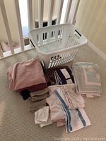 White plastic laundry basket and various folded and stacked bath and hand towels in different colors and patterns displayed on carpeted floor.