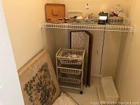 Wide view of wire shelf with sewing supplies, rolling cart, and vintage folding card table leaning against wall.