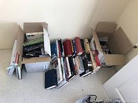 Three boxes filled with various hardback and paperback books arranged in rows on the floor.
