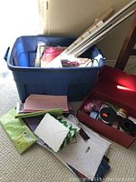 Large blue storage box filled with rolls of wrapping paper and tissue paper sheets, plus some envelopes and other wrapping accessories.