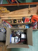 Open vintage wooden toolbox showing hand saw attached to lid, multiple compartments filled with hand tools such as pliers, measuring tapes, and screwdrivers. Black & Decker orange case visible in right compartment.