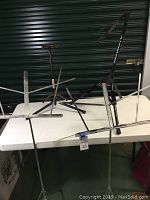 Two guitar stands, two metal music stands on a white table in front of green storage unit door.