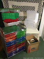 Full view of rolling cart with colorful drawers, plastic bins stacked on table, and cardboard box with office supplies on floor.