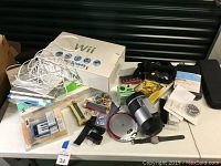 Nintendo Wii console in box along with Wii Fit balance board, several Wii remotes, power adapters, other gaming accessories, and miscellaneous small electronics and components on a white table.