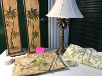 Two palm tree carved decorations behind a brass palm tree lamp with white shade, and a tropical leaf patterned tablecloth with matching coasters set on the table.