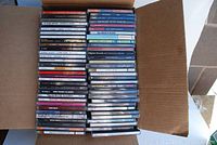 Front view of an open cardboard box with approximately 60 CDs arranged vertically, showing various artist and album titles on the spines.
