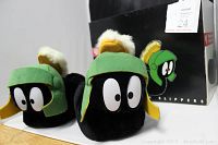 Two plush slippers shown from the front with Marvin Martian character design featuring large eyes, green helmet with yellow feather, black face and base.