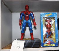 Photo of a vintage Spiderman bank figure standing on a gray brick style base next to a boxed Spiderman bobblehead with blue spider web themed box.