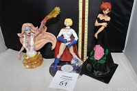 Photo showing both superhero figurines with a ruler for scale and auction tag 51. White outfit character with blast effect and anime character in black dress with rose graphic.