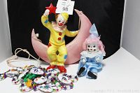 Front view of vintage ceramic clown on pink crescent moon with red star, seated pink-haired clown doll in blue satin, and Mardi Gras necklaces with masks and beads.