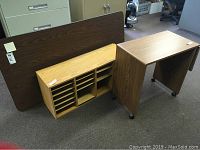Photo showing wood grain laminate folding table leaning against a filing cabinet, a wooden organizer shelf unit in front, and a wheeled typing cart on the right.