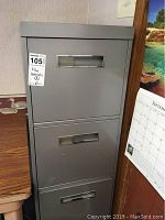 Grey 4-drawer steel file cabinet with stuck top drawer and tape on surfaces, placed next to wooden furniture and wall calendar