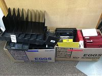 Two boxes containing multiple office organization items including black plastic document organizer trays, red desk trays, Webster's II dictionary, clipboards, and other desk tools.