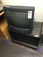 Sharp black CRT TV sitting on black media cabinet TV stand with wood-tone base and glass doors.
