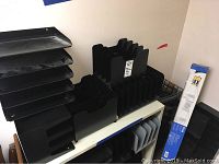 Black plastic letter organizer trays stacked on a shelf with black vertical file sorter racks beside them and black wire document baskets below.