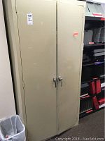 Closed beige steel storage cabinet with locking handles, showing some surface wear and tape on the doors. Cabinet next to black and red storage bins.