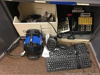 Photo showing Lucent multiline desk telephone, Dell KB212.B keyboard, Casio adding machine, and Powergear personal heater with user manual inside a cardboard storage box.