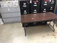Gray 3-drawer file cabinet and woodgrain folding table with black foldable legs on concrete floor.