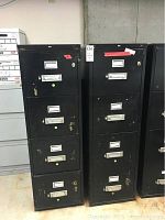 Two tall black fireproof file cabinets with multiple drawers and metal handles. Both have significant paint wear and scratches. One has a key lock, the other does not.