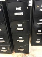 Front view of first black metal 4-drawer file cabinet with locks, handles, label holders, and key in lock, showing scratches and paint loss on surface.