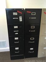 Frontal image of two black metal filing cabinets standing side by side, showing drawer fronts, label holders, handles, and surface condition including tape and scratches.