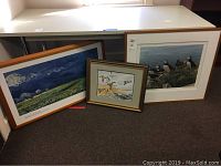 Three framed artworks side by side on carpet floor: large puffins print on right, Van Gogh poster on left leaning on table, smaller duck watercolor in middle