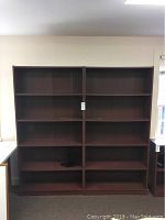 Two adjacent wooden bookcases each with four shelves, showing their full height and width, placed against a wall near a window and a desk.
