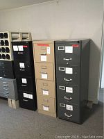 Photo shows three vertical four-drawer metal file cabinets in black, beige, and dark gray colors, positioned side by side against a wall, displaying labels on drawers and signs of wear.