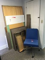 Photo showing parts of the disassembled drafting table including wooden tabletop, metal frame, and panels stacked upright against a wall with blue swivel chair next to them.