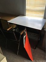 Drafting table with white flat top surface and black drafting chair positioned nearby, along with drafting pad attached to the table edge.