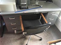 Full view of the metal desk with five drawers on left side, black office chair with wheels and armrest visible in front of the desk.