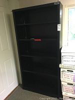 Tall black metal bookshelf with five shelves, placed against wall near window and door. Visible scratches and signs of wear on shelves.