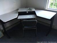L-shaped adjustable height computer desk with white top and black frame, gray padded chair in front, located near window
