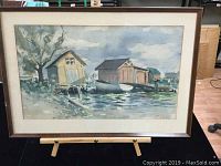 Full image of framed watercolor showing coastal scene with boathouses near water and trees.