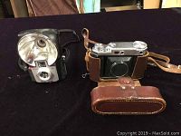 Two vintage Kodak cameras on a dark surface, Kodak Retina 2 folding camera in brown leather case with strap, Kodak Brownie Starflash box camera with large reflector flash.