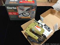 Green Black & Decker jigsaw inside open box, with power cord and instruction manual visible, alongside Clarke rotary tool in original box with accessory set box behind