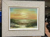 Framed oil painting on canvas depicting a sunset or sunrise seascape with birds, sea, and dune grasses.