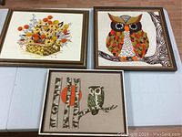 Set of three framed crewel embroidered hangings with owls and floral basket laid out on a table
