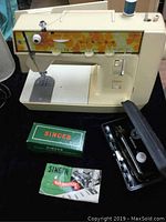 Front view of vintage Singer sewing machine with orange floral panel and accessories arranged in front including green box, buttonholer case, and instruction booklet