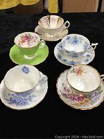 Photo showing the five cups and saucers with various floral patterns and gold trims on black background.