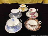 Five vintage bone china tea cups and saucers displayed on black fabric showing various floral and ornate designs.
