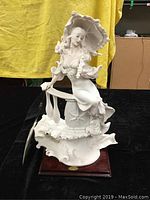 Full side view of the white resin figurine of a woman holding a parasol seated on a decorative base, against a yellow and black background.