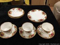 Photo showing 3 cups with matching saucers, 3 salad bowls, and 2 round plates in Old Country Roses pattern laid out on a black surface.